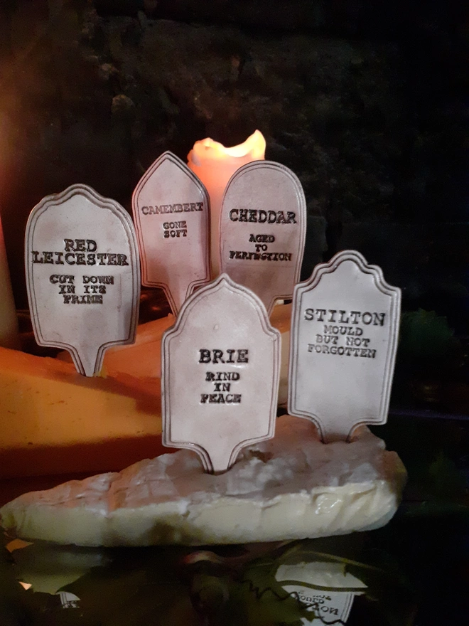 Close view of five handmade gravestone-shaped cheese markers with dark lettering and humorous cheese epitaphs, standing in wedges of cheese against a dark background.