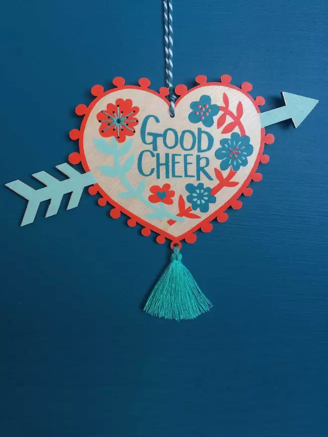 'Good Cheer' is hand printed on a floral wooden heart with an arrow through it. A tassel hangs down