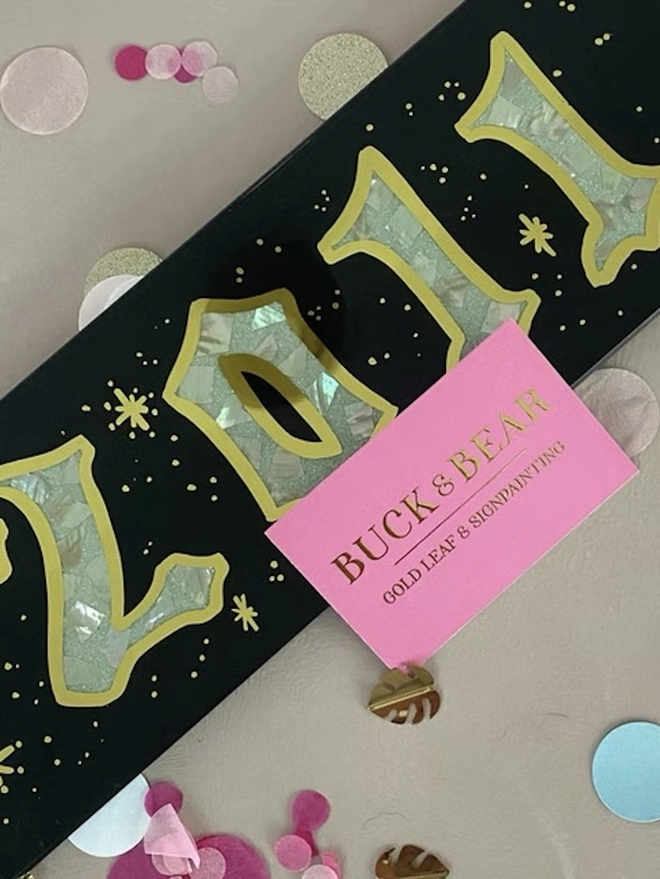 Personalised Year Date Sign, a black and gold personalised year sign sitting on a plain surface. It is joined by a small pink card and confetti. 