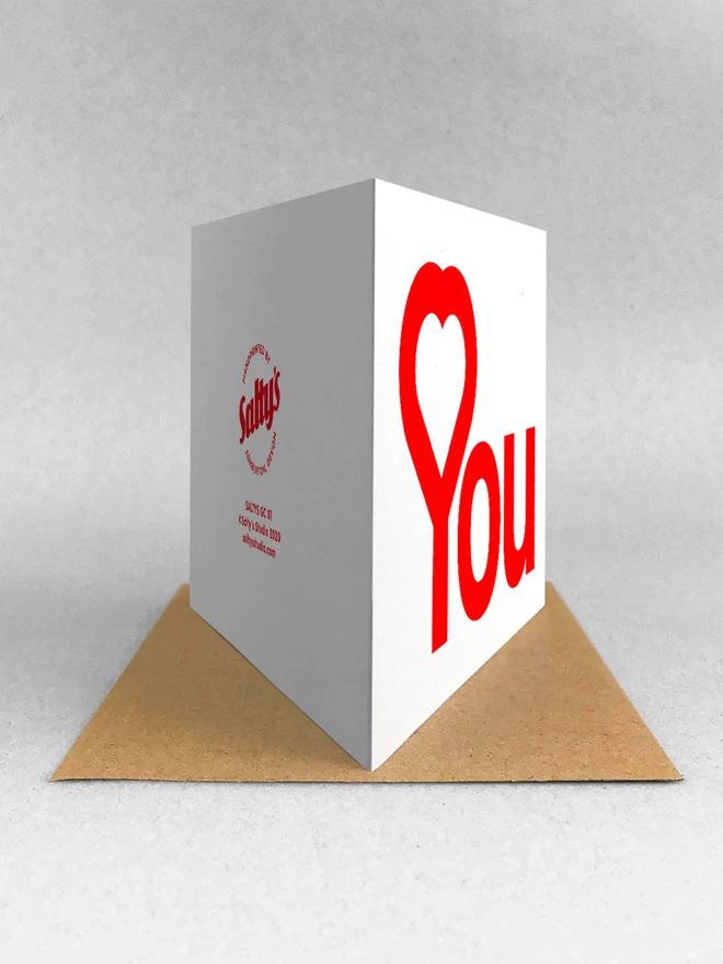 Rear view of a white card with the word You designed with a heart shaped into the top of the Y, reading Heart You. Stood on a brown Kraft envelope in a white backdrop, casting a light grey shadow.