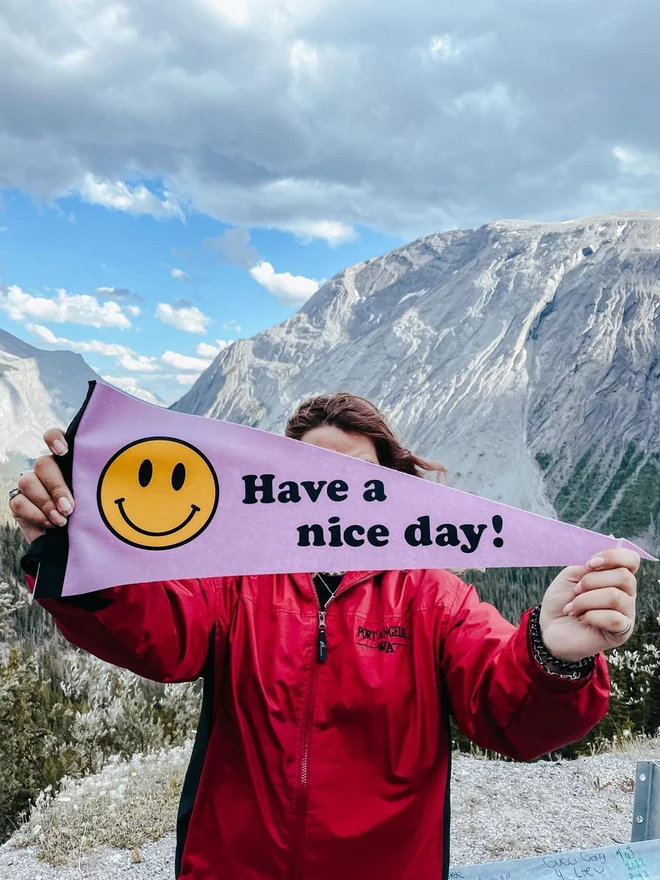 a girl hiker in a red jacket stands amid snowy mountains and a big sky, she is holding up a large felt pennant flag that is lilac with a smiley face on, and the words have a nice day!