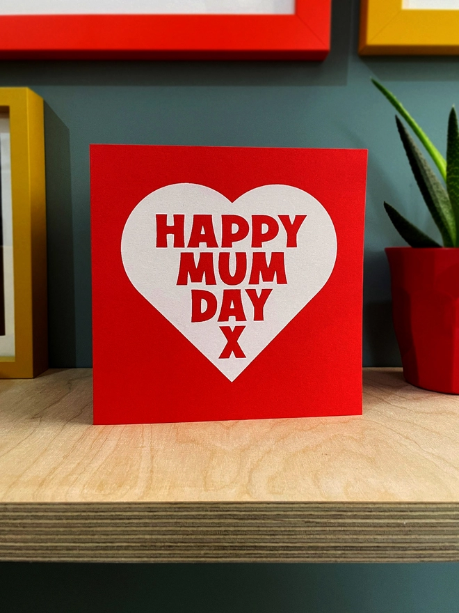 A red square card with a white heart, containing the words Mappy Mum Day x. Stood on a plywood shelf with hints of framed pictures around the edges and a plant half in shot.