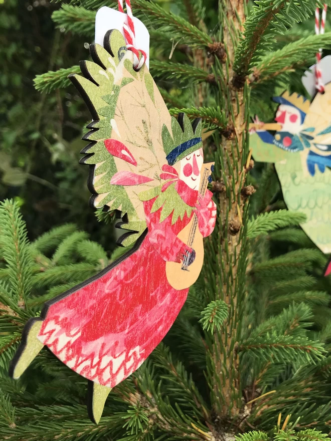 Red and green illustrated angel decorated on wood, designed by Esher Kent, with a Joyful Angel in the background, hanging against the fresh green of a fir tree Christmas tree
