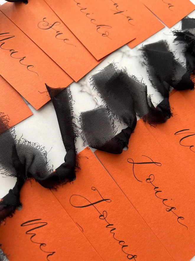 Personalised Calligraphy Rust Orange Place Card, orange place cards with black chiffon ribbons grouped together on a white surface. 
