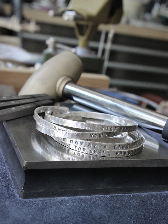A stack of sterling silver hammered cuff bangles resting on a metal block and a blue suede sandbag, in an jewellery workshop with steel punches and a brass mallet in the background. 