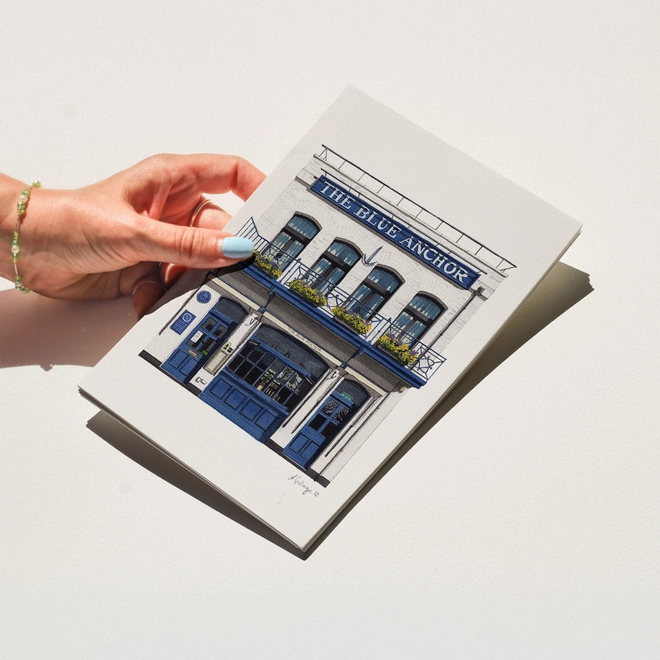 A hand with blue nail polish holding a print featuring an illustration of The Blue Anchor pub against a white background.