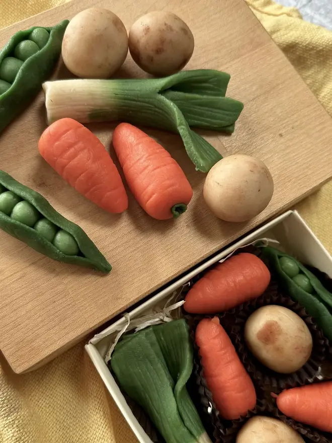 Veggilicious Marzipan Box, a marzipan vegetable box on a wooden chopping board on a yellow table cloth. In the box are marzipan peas, spring onions, potatoes and carrots. 