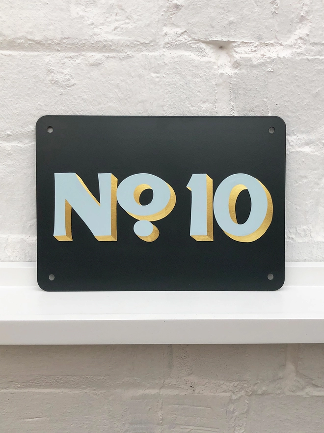 Hand painted house number No. 10 in pale blue and gold leaf on anthracite grey metal plaque, against a white brick wall. 