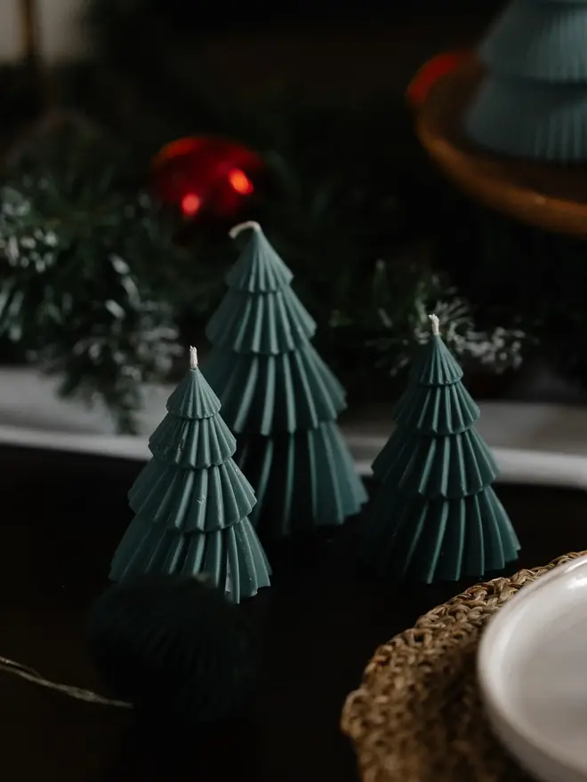 Forest Green Christmas Tree Warm Candles (Set Of 2). Three unlit deep green Christmas tree candles in graduating sizes on a dark table, with frosted greenery and a red bauble behind. The fluted sculptural design makes an elegant festive gift or table decoration.