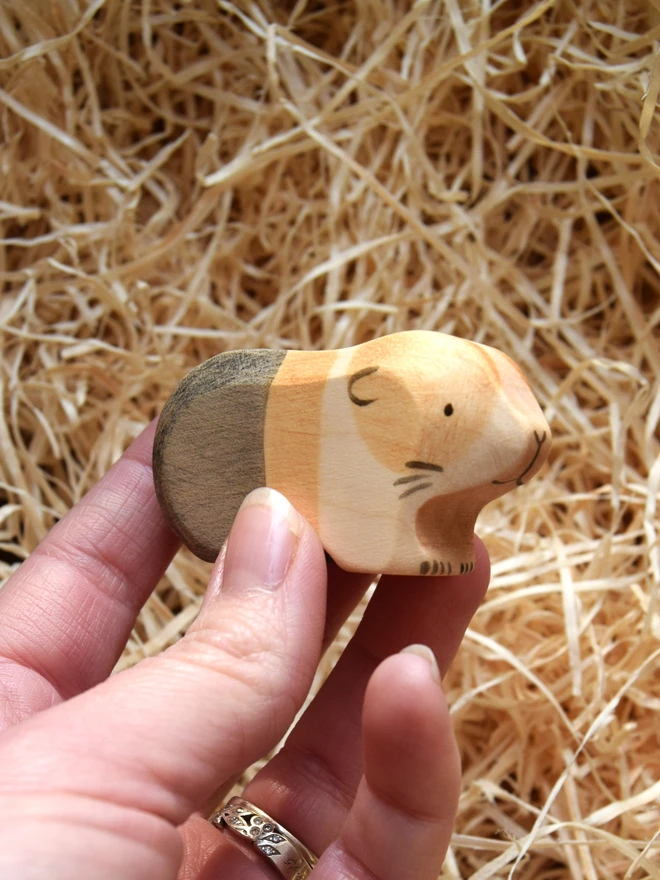 tri coloured qooden toy guinea pig held above woodwool