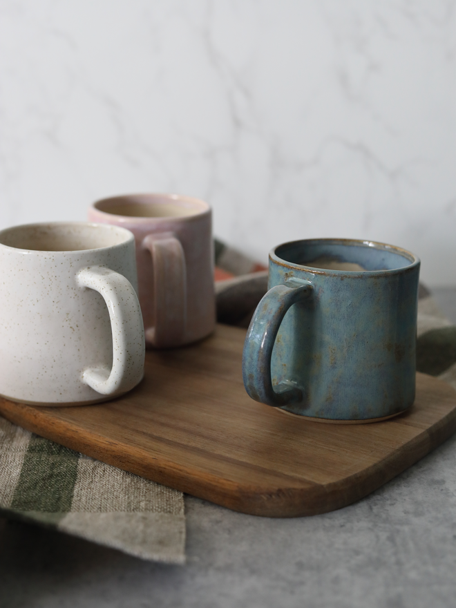 Three handmade ceramic standard mugs on a wooden board — speckled white and blush pink in the foreground, with a teal glaze mug alongside, each containing a hot drink, styled with a green checked linen cloth against a white marble background