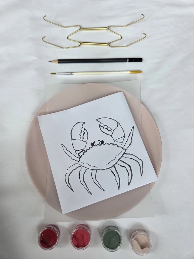 Contents of the crab plate painting kit on a white cloth - plate, design template, brush, pencil, paints and wall hook. 