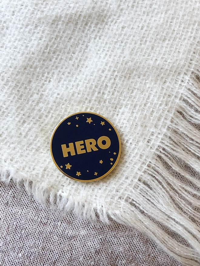 A navy blue and gold enamel pin badge with a starry design and the word "Hero" is pinned to a white scarf.