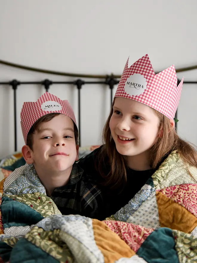 Two children wearing red gingham personalised Christmas crowns are wrapped in a cosy patchwork quilt giggling together.