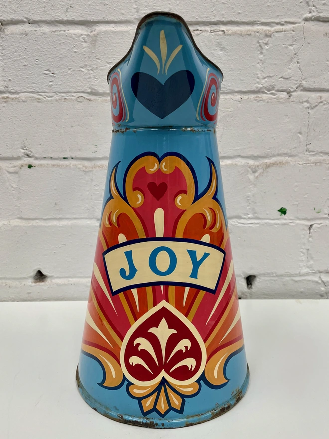 Large blue enamel jug hand-painted with a traditional fairground inspired design, in pink, orange and yellow. 'Joy' is painted in a banner across the centre.