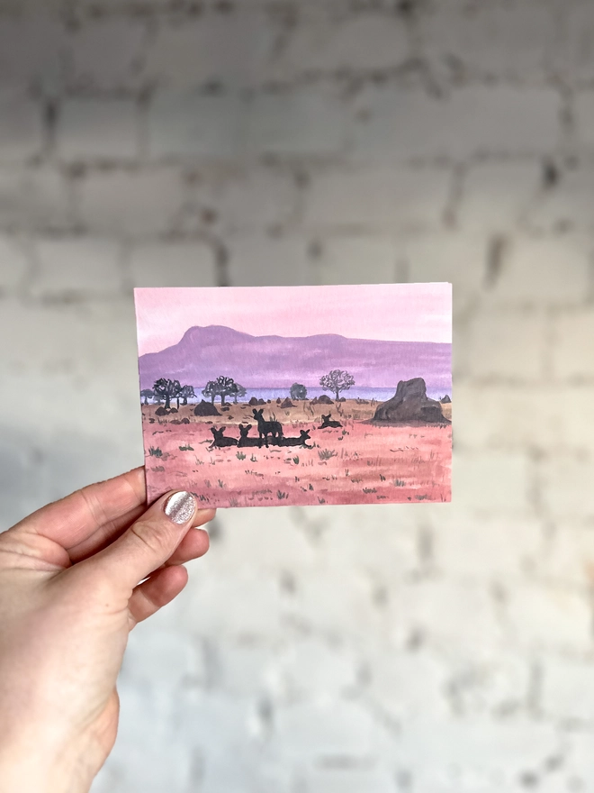 a greetings card featuring an illustration of a dusky coloured landscape and the silhouettes of some african wild dogs