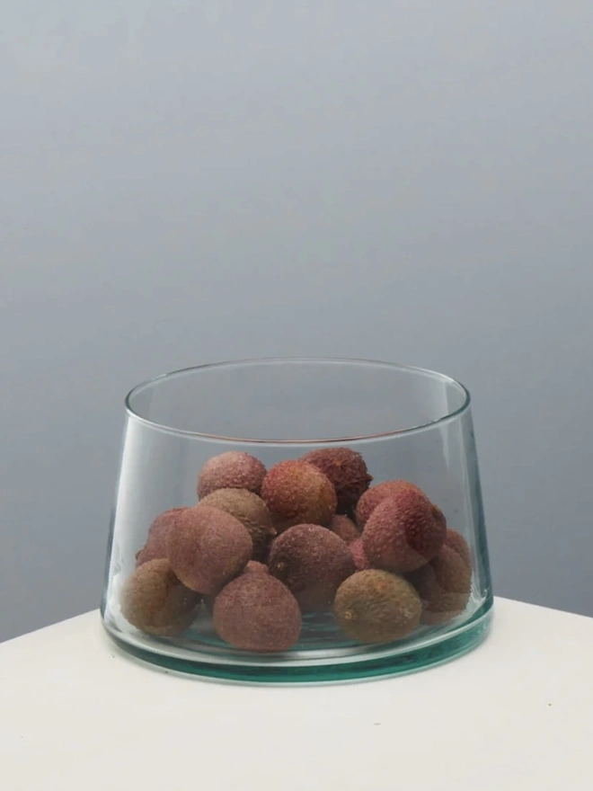 Tall Glass Dish, a tall glass dish sitting on a white surface against a baby blue backdrop. It is filled by lychees. 