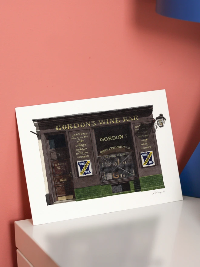 A framed watercolor print of Gordon's Wine Bar sitting on a white mantelpiece against a pink wall.