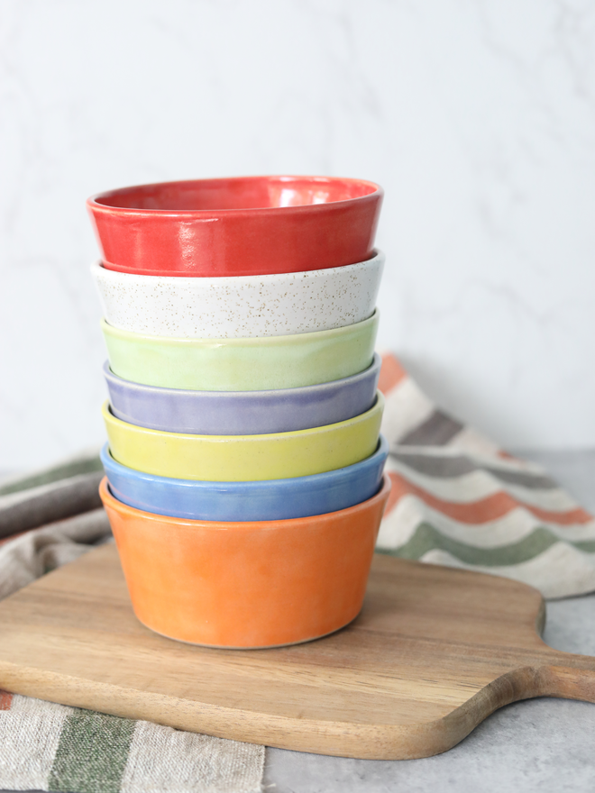 A neatly stacked set of seven glazed ceramic breakfast bowls in rainbow colours — red, speckled white, green, blue, orange, yellow, and purple — on a wooden chopping board, surrounded by linen napkins, against a pale grey background.