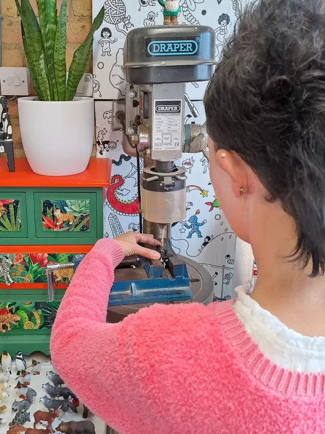 A hand holds a plastic and metal space shuttle on a clamp on the table of a pillar drill. A white tray of plastic animals sits below the pillar drill to the left. There is a small chest of drawers decorated in a brightly coloured jungle theme with animal drawer knobs attached with a snake plant in a white vase sitting on top. The space shuttle is being drilled and made into an space themed light switch by Candy Queen Designs.