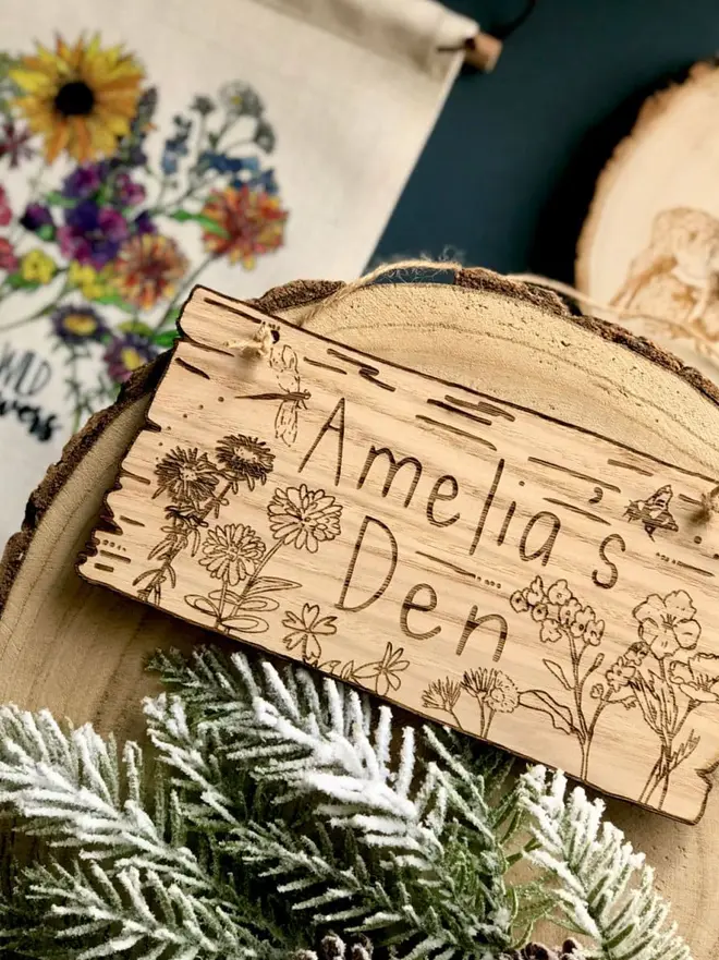Wild Flowers Den Sign, a wooden sign reading "Amelia's Den" with wildflower engravings sits on a tree slice next to a floral bag and snowy pine needles.