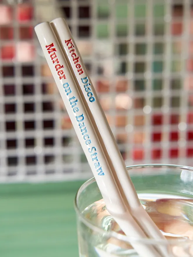 Pair of glossy ceramic cocktail straws with playful disco-themed text, photographed in a clear tumbler with soft-focus mosaic tiles behind.