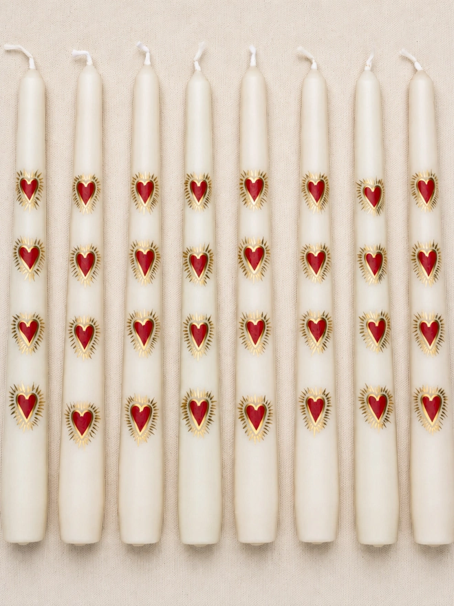 Close-up of ivory hand-painted taper candles arranged in neat rows on cream linen, each decorated with repeating red sacred hearts outlined in metallic gold.