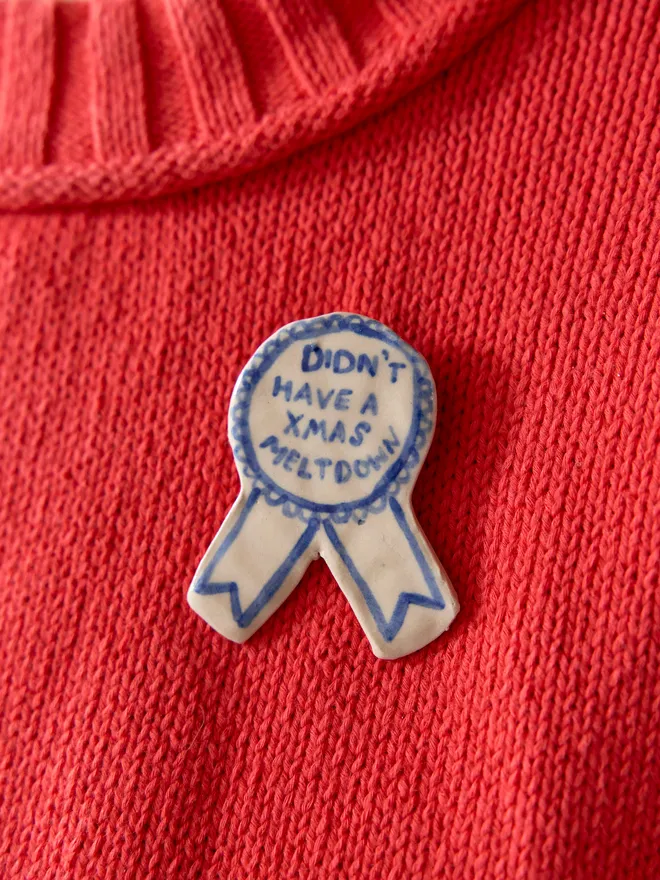 'Didn't have a Xmas Meltdown' hand-painted on a ceramic Christmas award badge in blue, pinned to a woolen red knitted jumper
