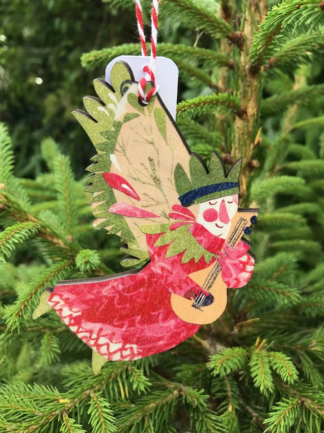 Red and green illustrated angel decorated on wood, designed by Esher Kent, hanging against the fresh green of a fir tree Christmas tree