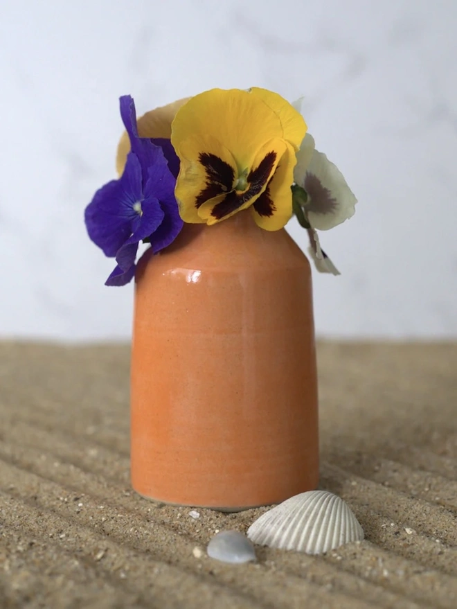 Classic Bud Vase, a colourful ceramic vase sitting on a plain surface against a grey backdrop. It is accompanied by some flowers and shells. 