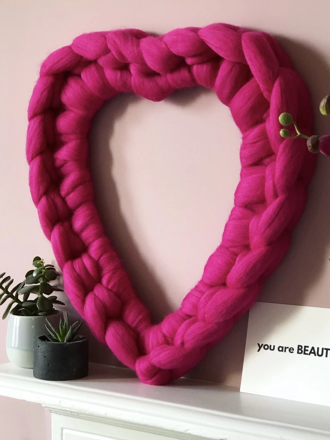 A bright pink woolly heart wreath sitting on a white mantel