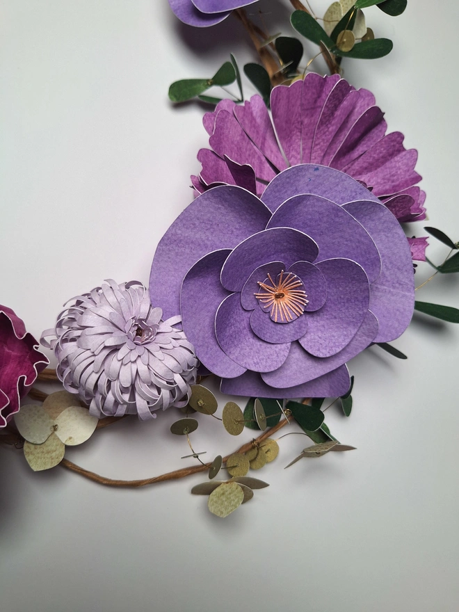 Close up of purple paper flowers, depicting various species in different shades of purple. The central flower in this picture is a camelia, with zinnia and chrysanthemum flowers nestled beside it. 