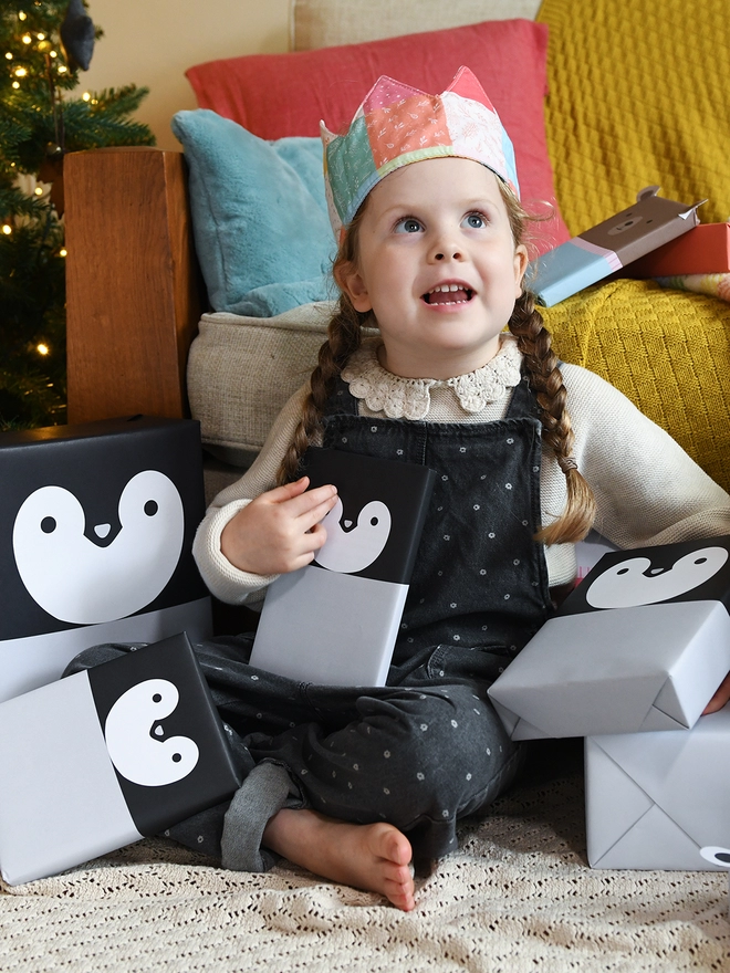 A young girl wearing dungarees and a patchwork crown is holding several gifts wrapped as penguins.
