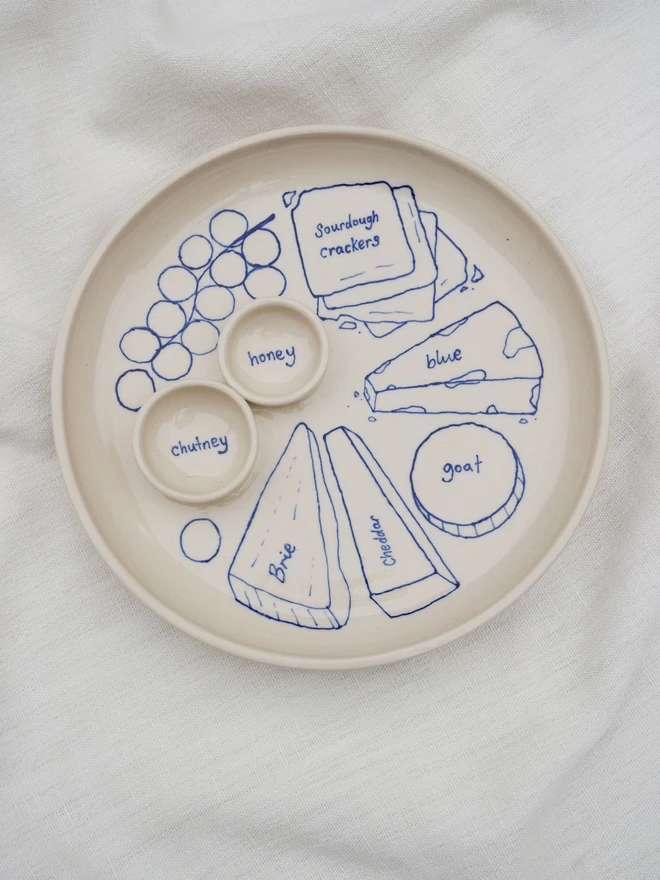 Dippy Cheeseboard Dinner Plate, a white dip plate sitting on a bed cloth. It's covered in blue outlines of cheeses, crackers and grapes.