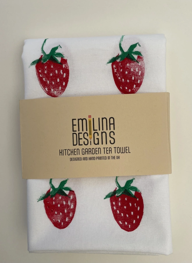 Strawberry print kitchen tea towel folded on a wooden chip table with a cardboard packaging band.