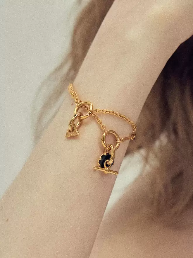 Woman wearing two gold bracelets styled with a triangle charm, a black onyx disc and a mini Albert lock charm