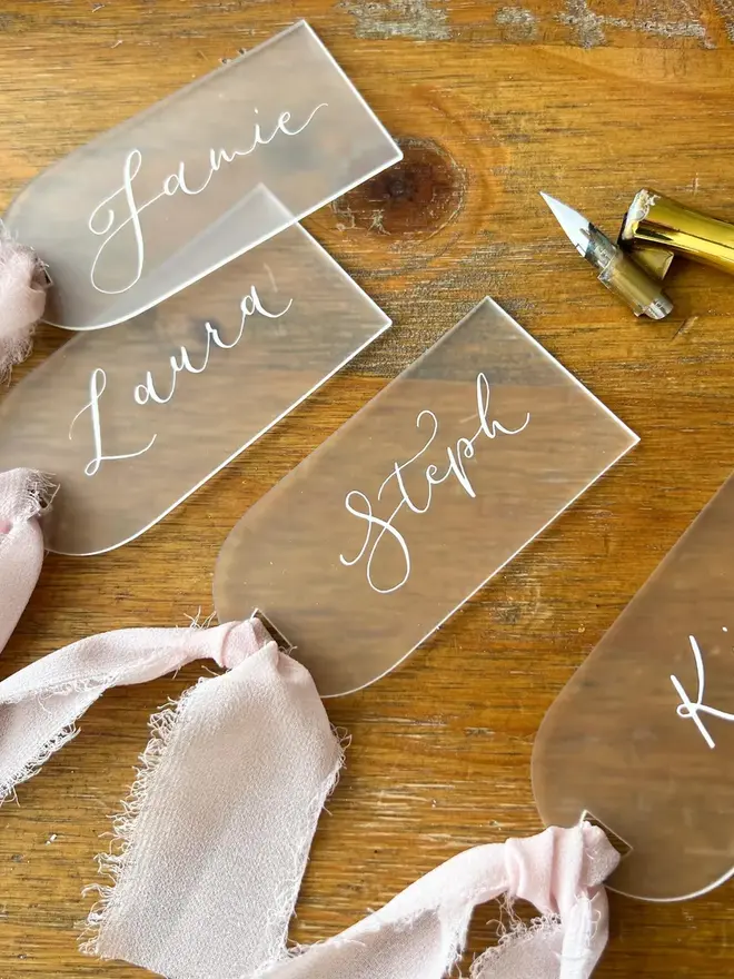 Personalised Calligraphy Frosted Acrylic Place Cards, frosted acrylic place cards with pink ribbons grouped together on a wooden table. 