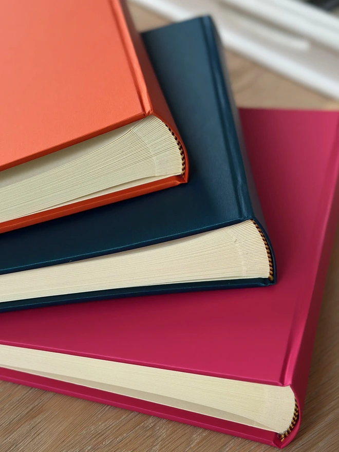 A pile of hardback photo albums rests on a wooden surface. The colours are orange, blue and pink.