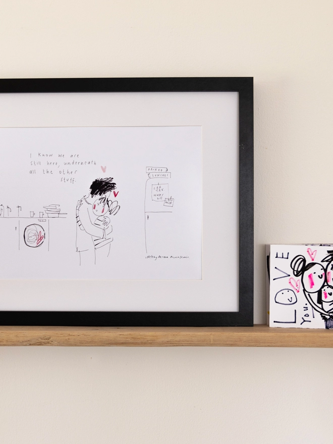 A sketchy muma print in a black frame on a shelf next to a black and white illustrated card. A line drawing print of a man an women hugging in a domestic setting.