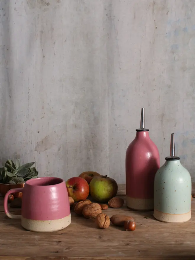 Ceramic Fuchsia mug sat on a wooden surface. The background shows our large fuchsia oil pourer and a small mint green oil pourer sat amongst scattered seasonal fruits.
