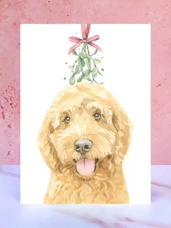 A Christmas card featuring a hand painted design of a Ruby Cockerpoo, stood upright on a marble surface.