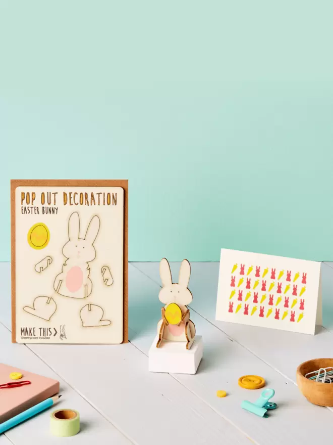 3D Easter bunny decoration and rabbit and carrot pattern greeting card and brown kraft envelope on top of a grey desk in front of a light green coloured background