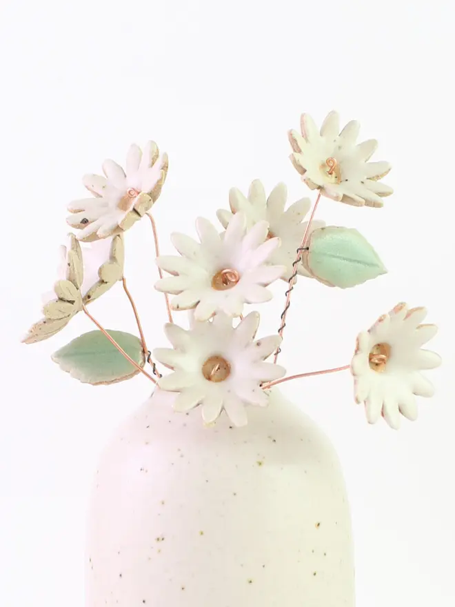 Eight ceramic large white daisies with copper stems and two mint green ceramic leaves coming out of a speckled white ceramic vase