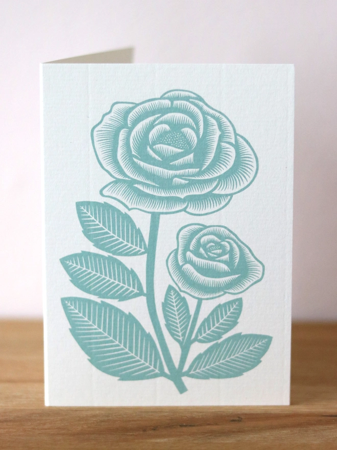 A greetings card feature a teal rose design based on an original linocut print