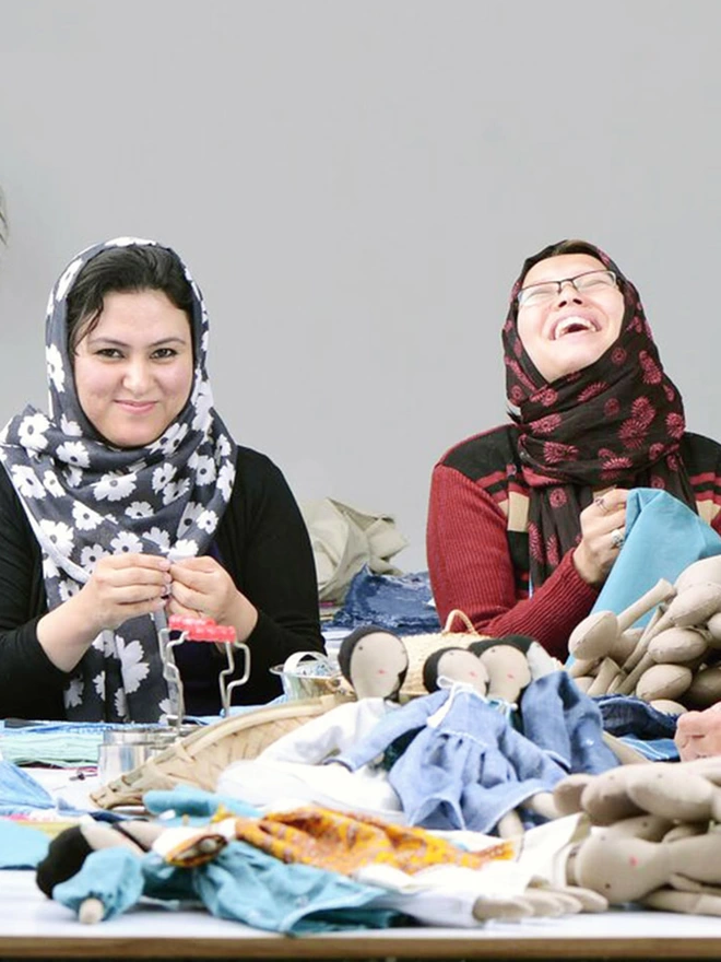 Tow ladies laughing and stitching fabric next to a pile of fabric dolls ready to be dressed