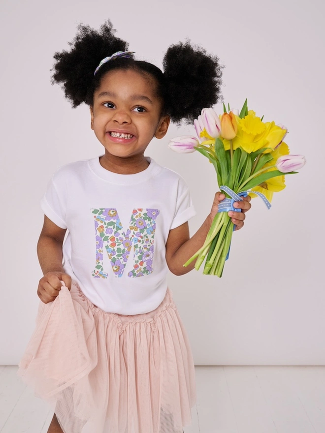 a 3 year old girl wearing a personalised kids white t-shirt with an initial cut from betsy purple liberty floral print on the front