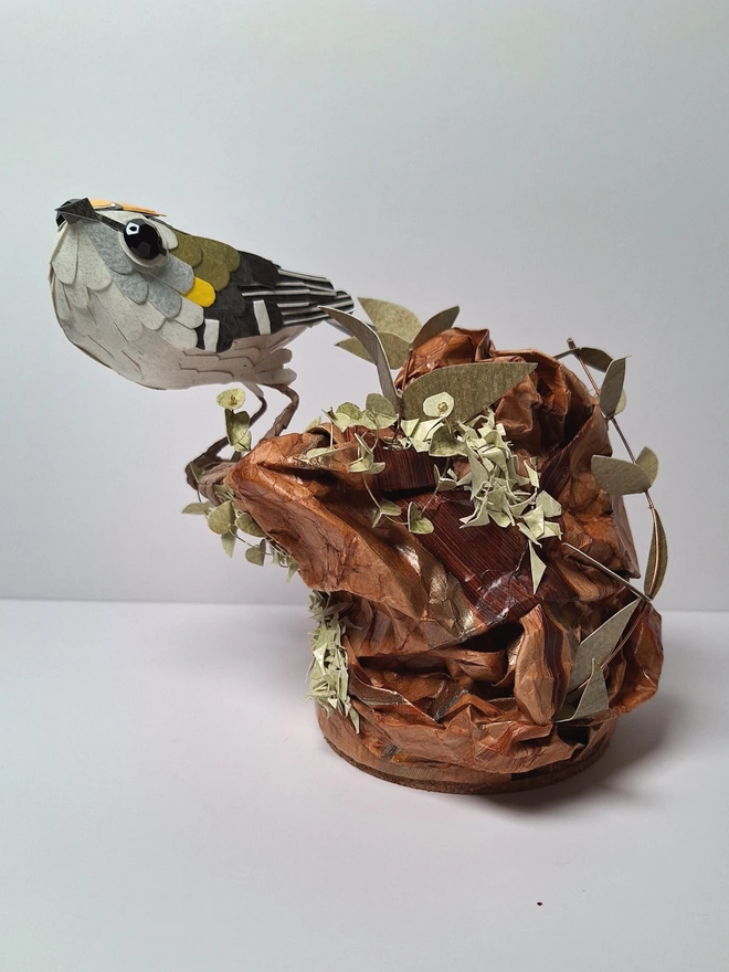 Firecrest paper bird sculpture, perched on a tree stump. Front view.