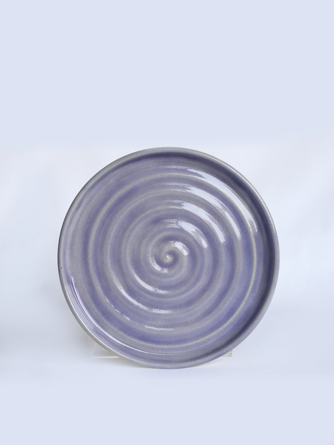 A handmade ceramic cake plate in amethyst purple glaze, photographed from the front against a plain light grey background, showing a natural concentric swirl texture and a shallow raised rim