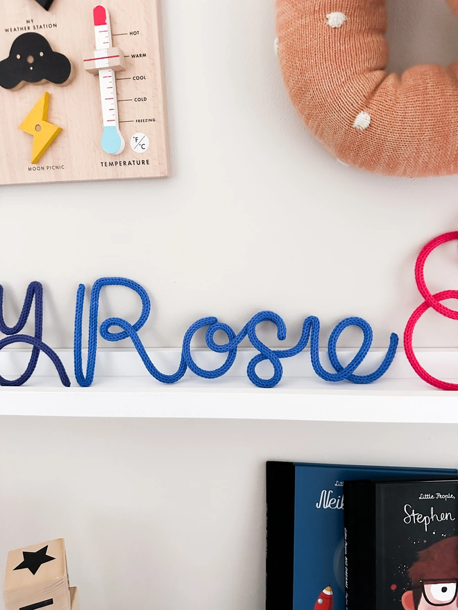Personalised Baby Name Sign 'Rosie' on a white shelf in a child's bedroom with other kids decor and childrens books displayed around it. 
