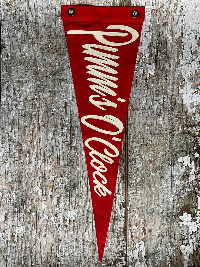 A red canvas pennant flag with Pimm's O'Clock written in ivory canvas hanging vertically on a wall. 